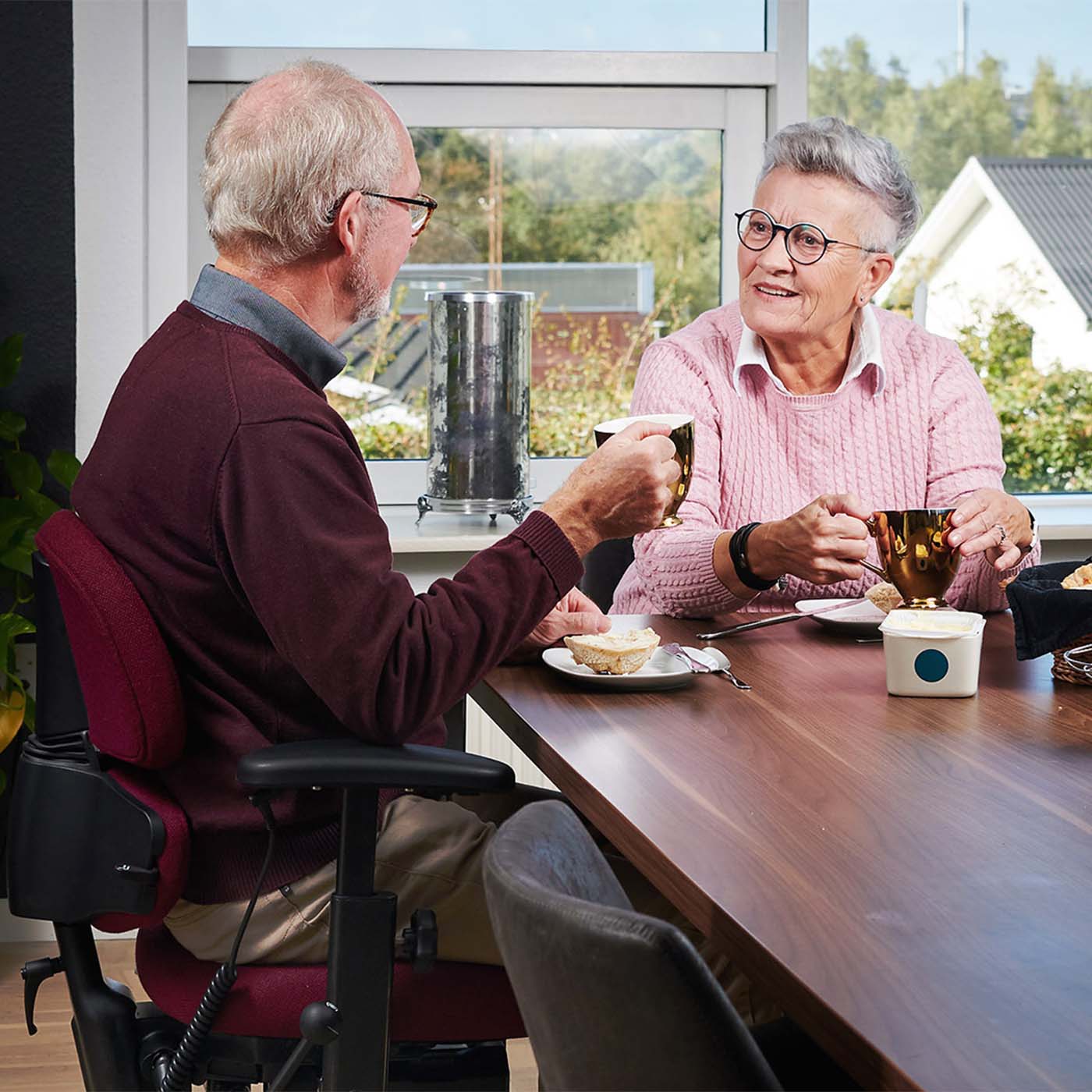 Zwei Senioren unterhalten sich am Esstisch. Einer sitzt im Bürostuhl mit elektrischer Höhenverstellung.