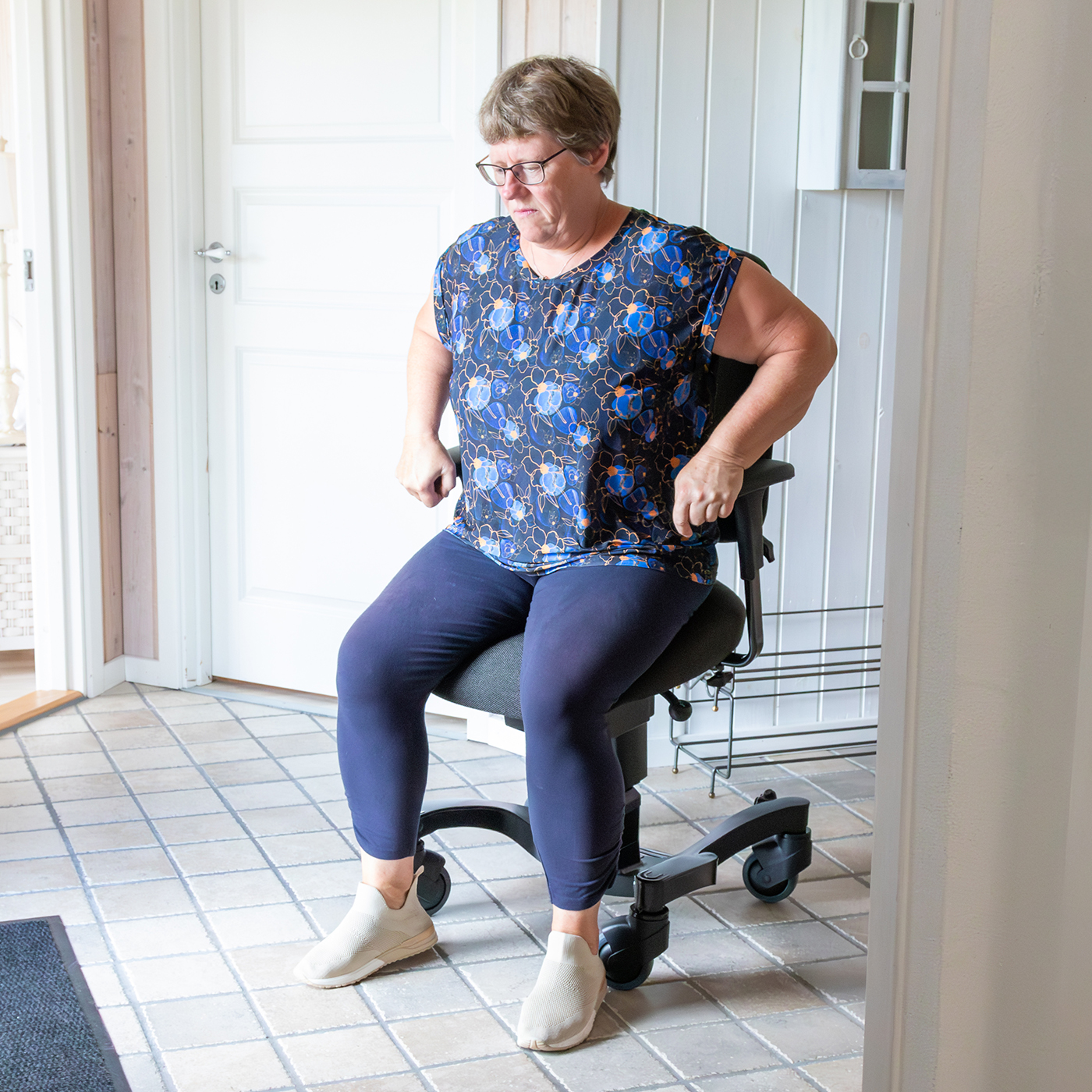 Frau nutzt eine Aufstehhilfe vom Boden in Form eines mobilen Stuhls, um sicher und selbstständig im Flur aufzustehen und sich fortzubewegen.