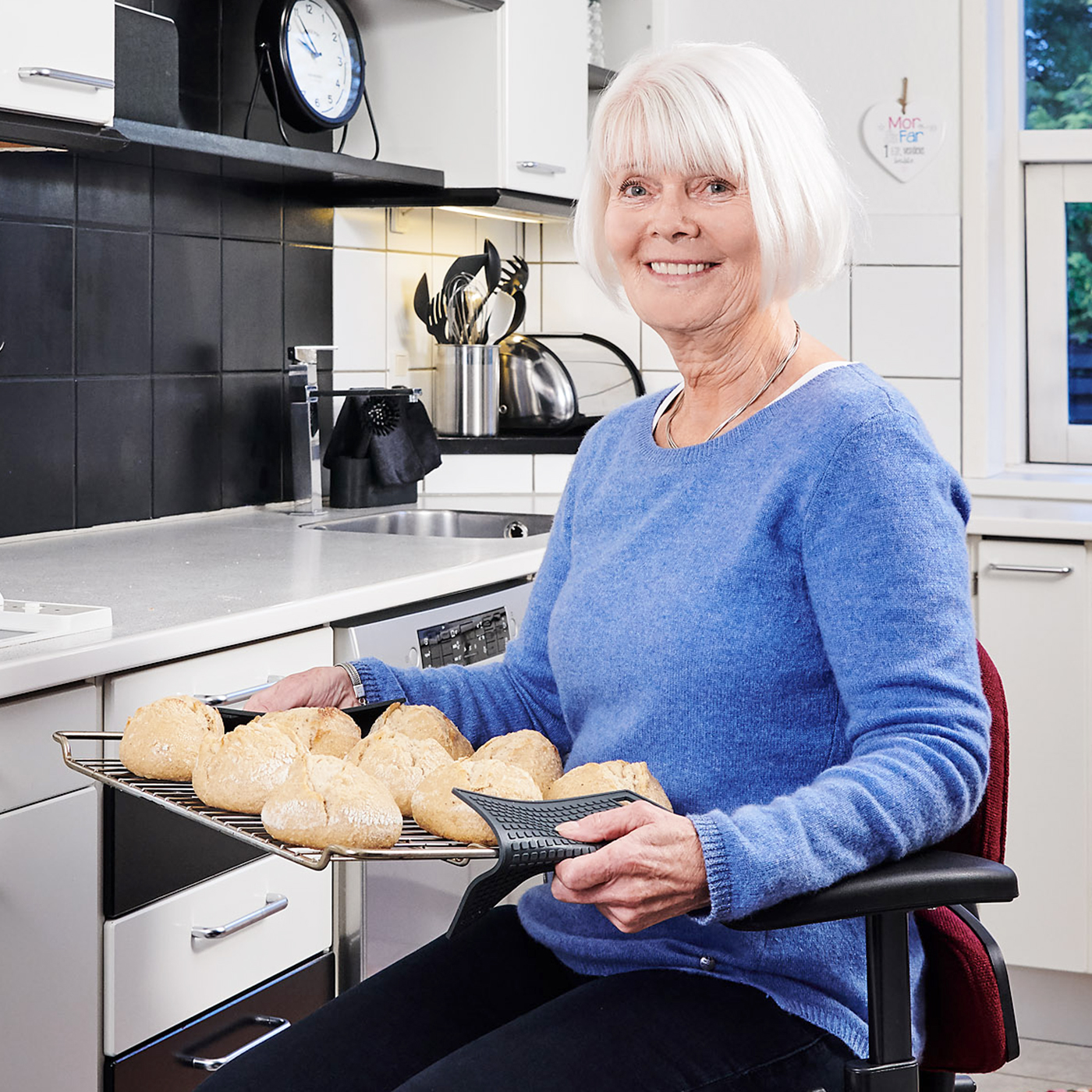 Seniorin sitzt auf einem Trippelstuhl in der Küche und hält ein Backblech mit Brötchen. Ein Trippelstuhl kann Menschen mit eingeschränkter Mobilität bei alltäglichen Aufgaben unterstützen.